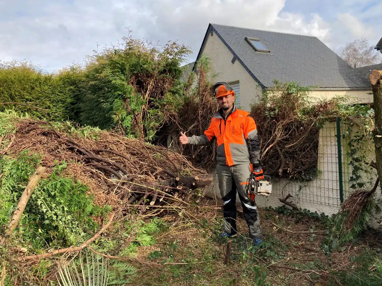Élagueur STIHL satisfait montre pouce levé près d'un tas de branches de thuya coupées devant une maison.