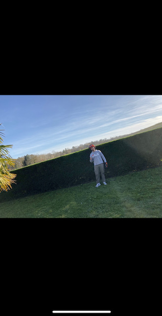 Homme portant un sweat-shirt FILA gris et une casquette rouge pose en faisant un signe de victoire sur une pelouse verte devant une haute haie taillée sous un ciel bleu clair.